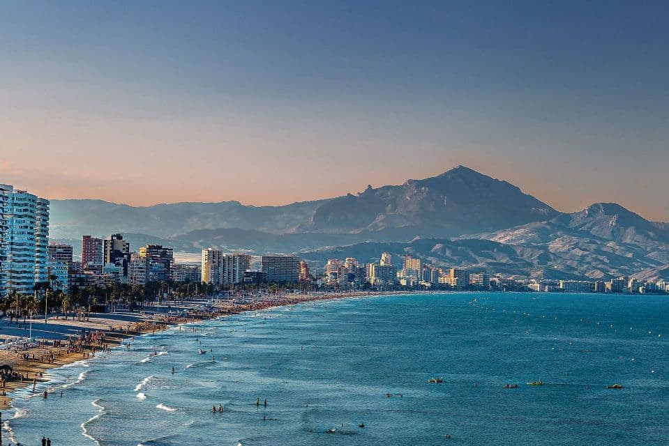 Una vista aerea di una spiaggia affollata lungo una città costiera, con una grande catena montuosa sullo sfondo sotto un cielo limpido.