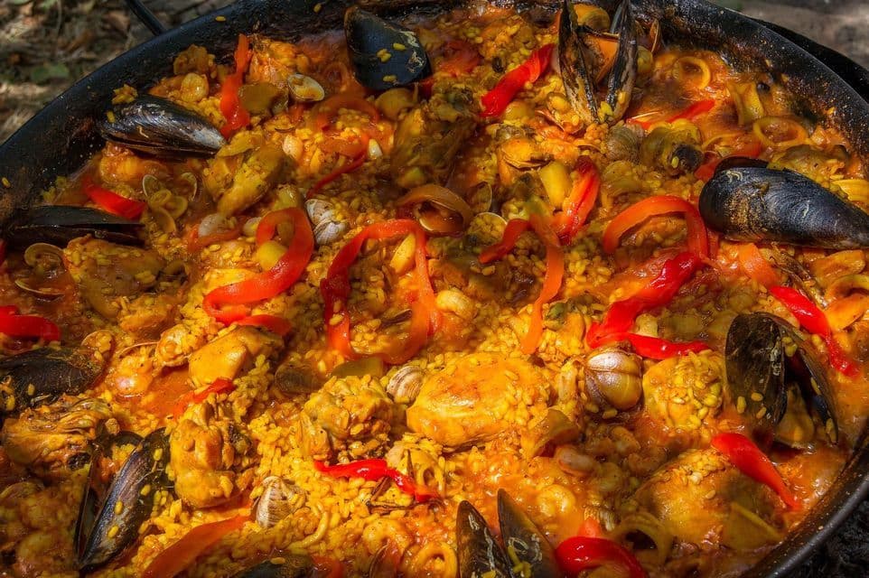 Un primo piano di una paella tradizionale in cottura in una grande padella nera, ricca di riso, pollo, cozze e peperoni rossi.