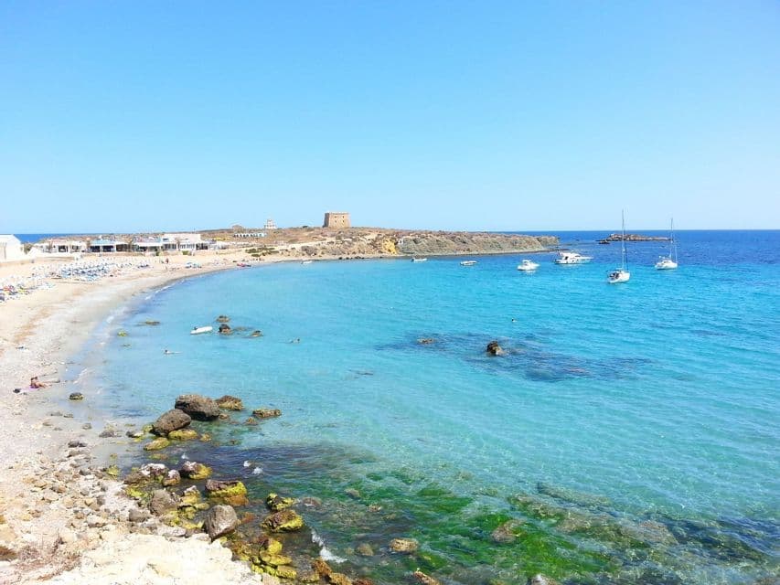 Una baia costiera soleggiata con una spiaggia sabbiosa, acqua cristallina turchese, diverse imbarcazioni e una torre di pietra su un promontorio lontano.