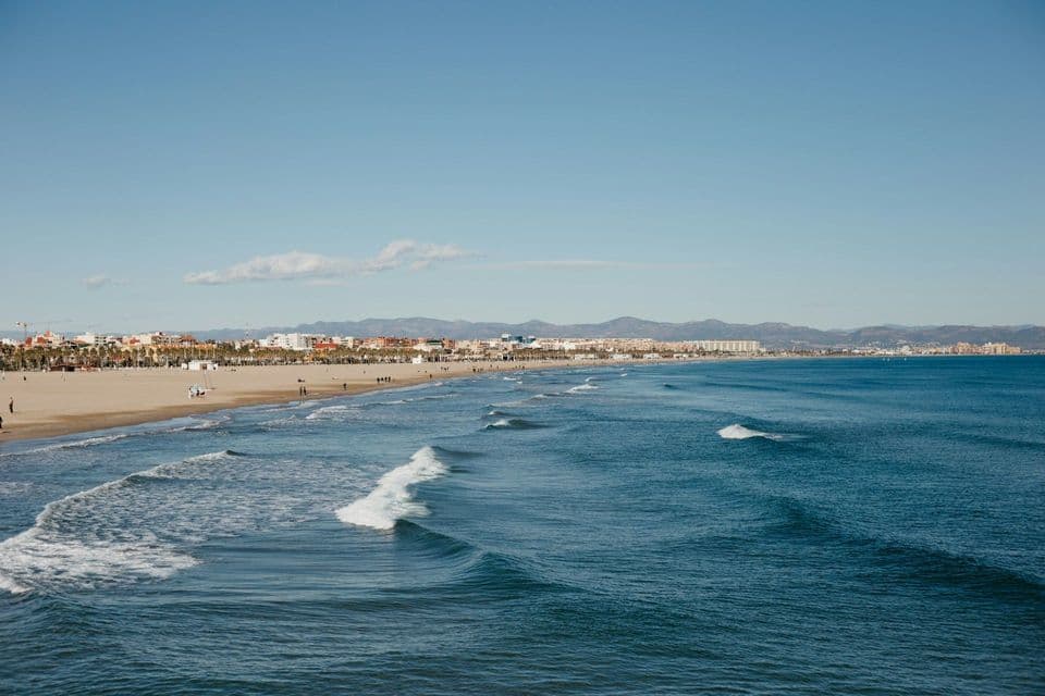 Le onde si infrangono su un'ampia spiaggia sabbiosa delimitata da una città costiera e montagne lontane sotto un cielo azzurro e limpido.