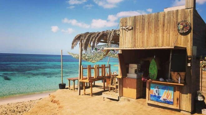 Un chiringuito rústico de madera con sillas apiladas se encuentra en una orilla arenosa, con vistas a un tranquilo océano turquesa bajo un cielo azul.