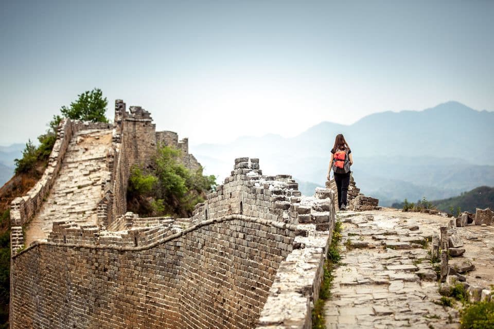 Una mujer con mochila camina por la cima de un muro de piedra desmoronándose que se extiende hacia montañas lejanas y brumosas.
