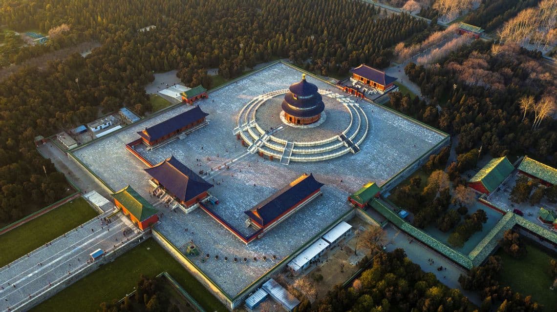 Una vista aérea de un complejo de templo tradicional chino con una pagoda circular de techo azul en una gran plaza rodeada por un denso bosque.