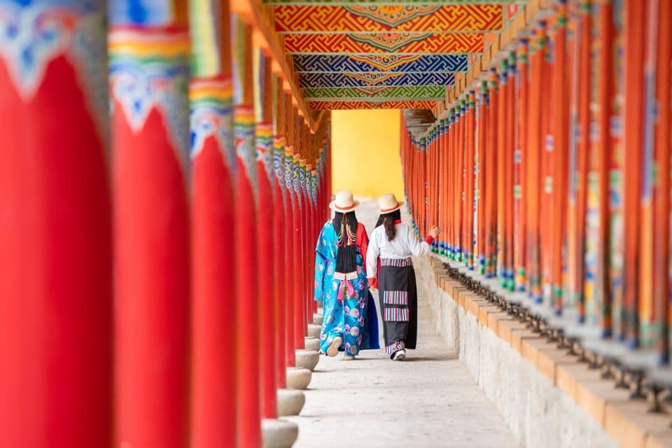 Dos mujeres con vestimenta tradicional y sombreros caminan por un corredor de templo bordeado de pilares rojos y ruedas de oración.
