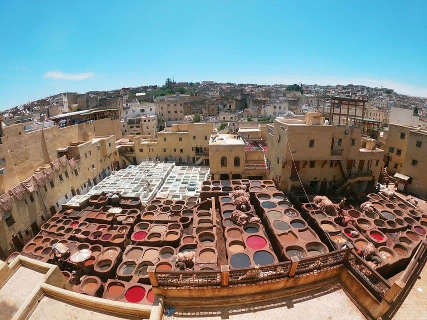 Veduta a fisheye dall'alto di una conceria tradizionale con numerose vasche in pietra contenenti tinture colorate, immersa in un fitto paesaggio urbano.