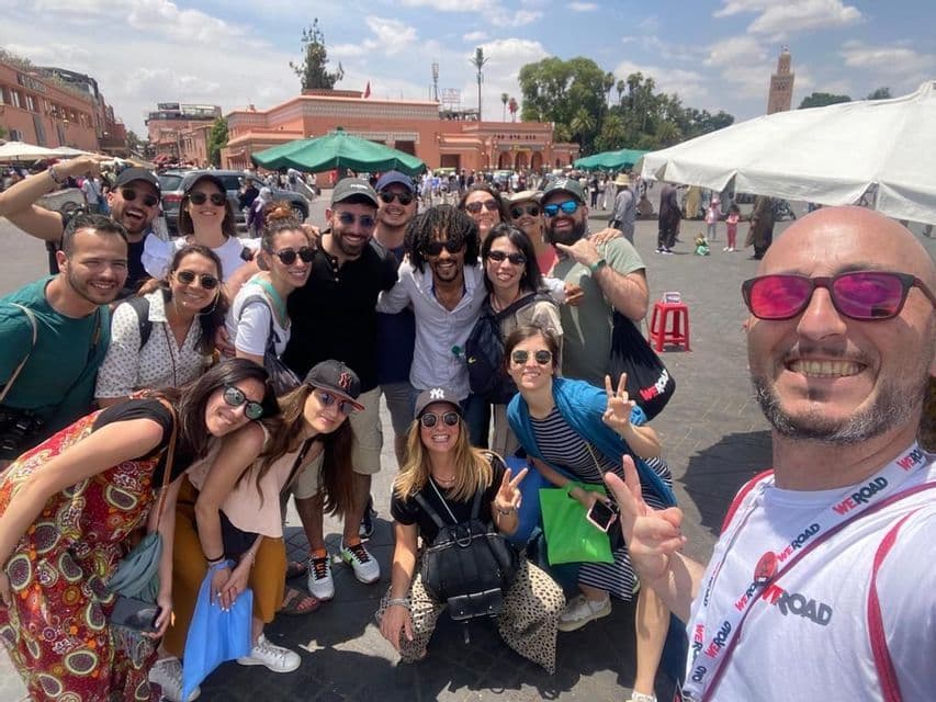 Un grupo grande de WeRoad sonríe y posa para una selfie en una bulliciosa plaza de la ciudad en un día soleado.
