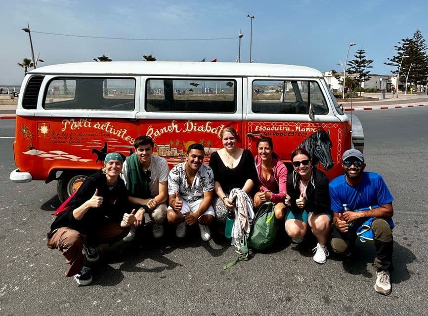 Un groupe WeRoad souriant et accroupi pour une photo devant un van vintage rouge et blanc sur une rue pavée.