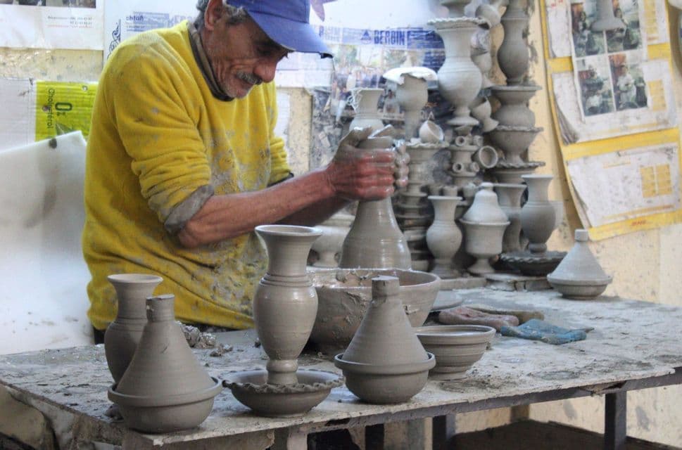 Un artesano con un suéter amarillo y gorra azul moldea un jarrón de arcilla en un torno de alfarero, rodeado de cerámica terminada en un taller.