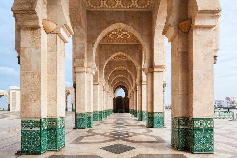 Una vista de una larga columnata de pilares de piedra y arcos moriscos, con detalles de azulejos verdes y un suelo estampado.