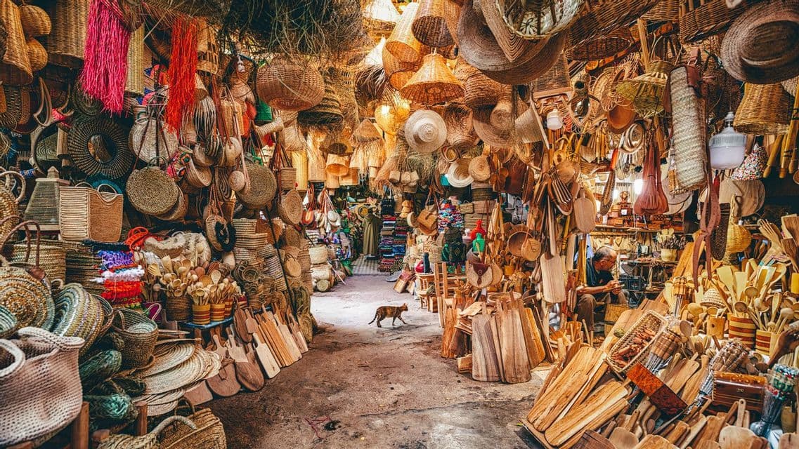 Un gato camina por el pasillo de un estrecho puesto de mercado abarrotado de cestas tejidas, sombreros de paja y artesanías de madera.