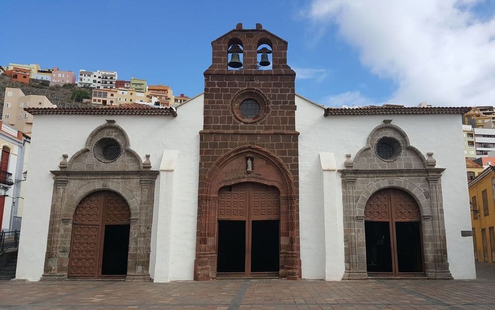 La facciata di una chiesa bianca con un campanile centrale in pietra marrone e tre portali ad arco, su una piazza pavimentata.