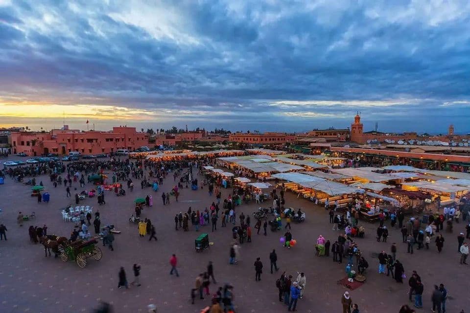 Una vista aerea di una vivace piazza cittadina, affollata di persone e bancarelle illuminate, al tramonto sotto un cielo nuvoloso.