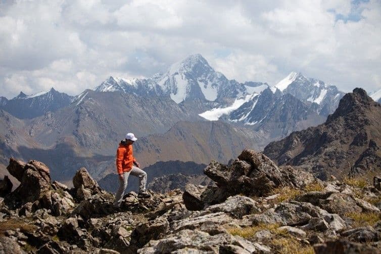Una persona con una giacca arancione fa un'escursione lungo un sentiero roccioso di montagna con cime innevate in lontananza sotto un cielo nuvoloso.