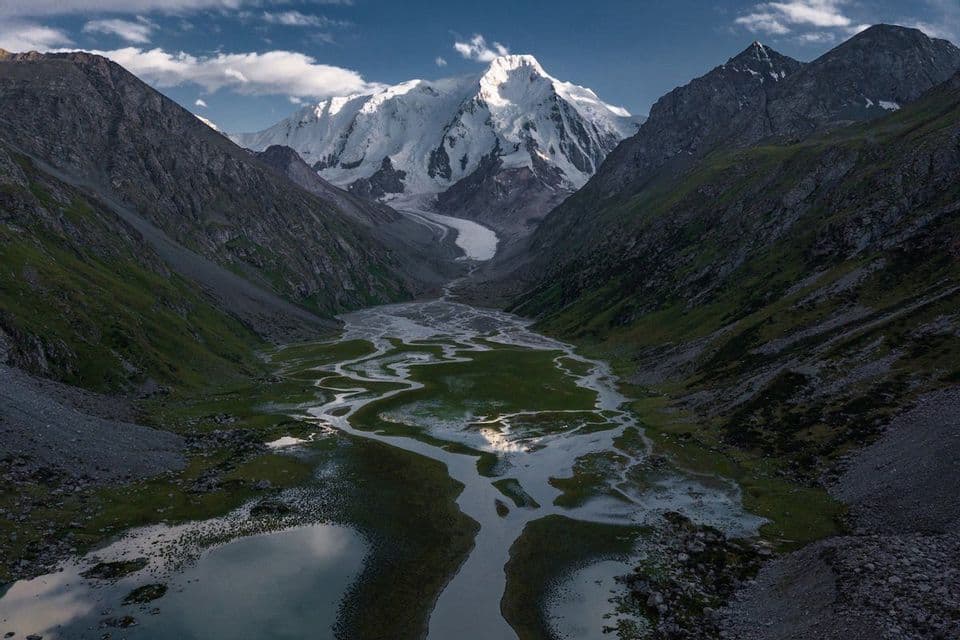 A braided river runs through a wide green valley, with a large snow-covered mountain and glacier in the background under a blue sky.