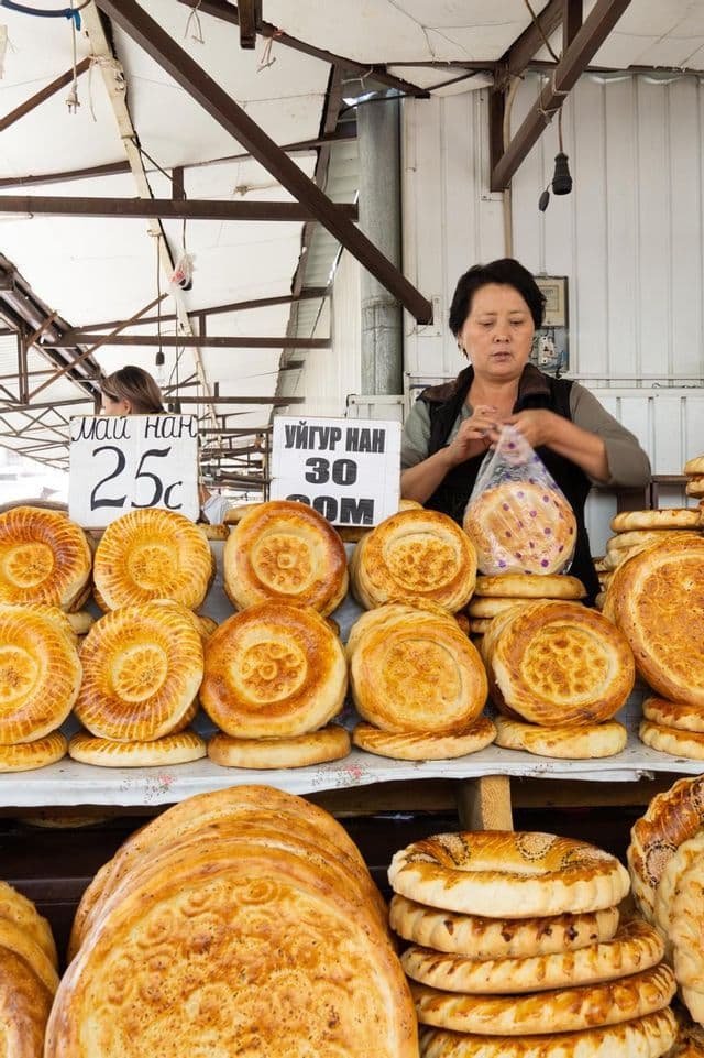 Une femme, à un étal de marché en plein air, dépose un grand pain plat, rond et décoré dans un sac en plastique, d'autres pains étant empilés et prêts à la vente.