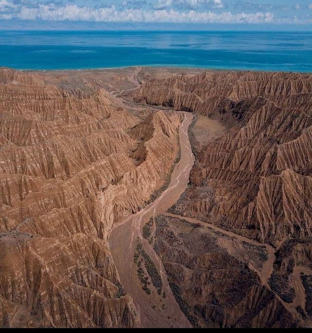 An aerial view of a winding river flowing through a large, eroded brown canyon towards the sea under a cloudy sky.
