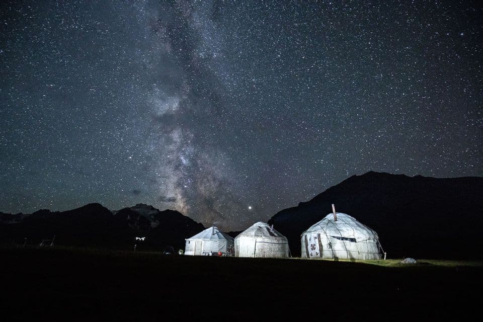 Trois yourtes illuminées se trouvent dans un champ la nuit sous un ciel étoilé montrant la Voie lactée, avec des montagnes en arrière-plan.