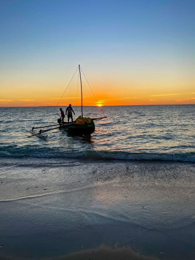 Dos personas de pie en un pequeño velero con balancín en el mar, silueteadas contra un atardecer dorado en el horizonte.