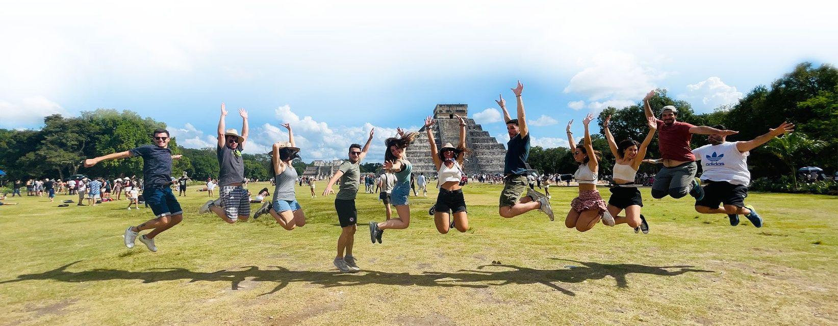 A WeRoad group trip of people jumping together on a grassy field with a large stone pyramid in the background.