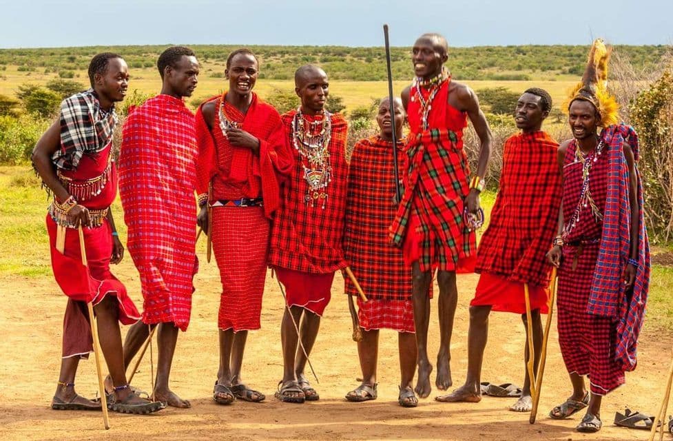 Un grupo de hombres masái con shukas tradicionales de cuadros rojos y joyas de cuentas, posan sonriendo en un camino de tierra en la sabana.