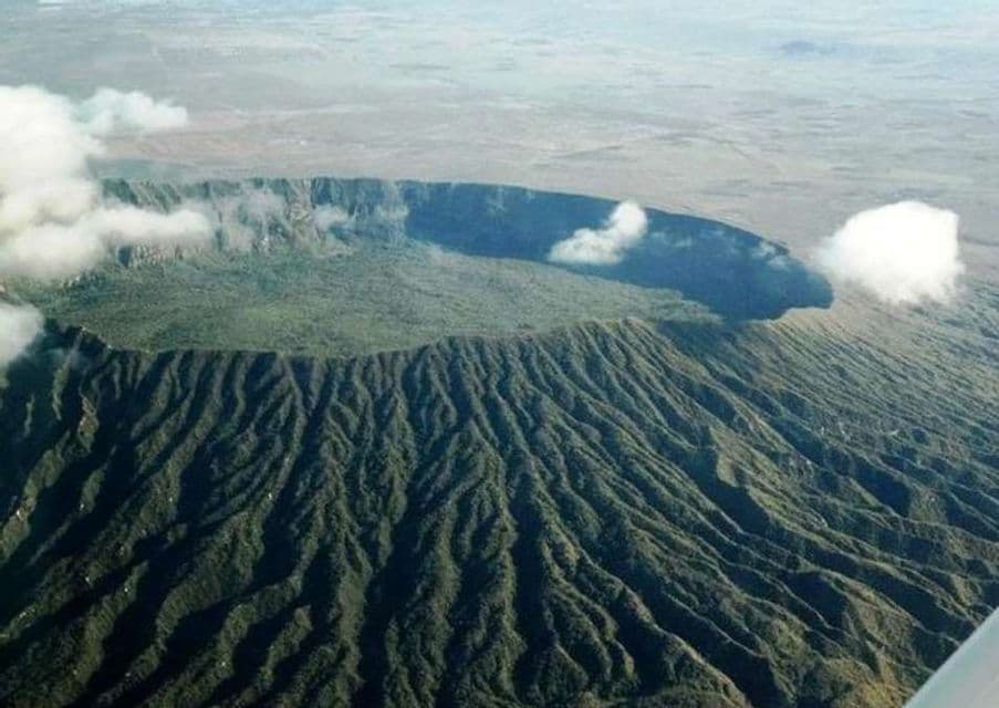 Una vista aérea de un gran cráter volcánico, sus laderas erosionadas y caldera cubiertas de vegetación verde bajo unas pocas nubes.