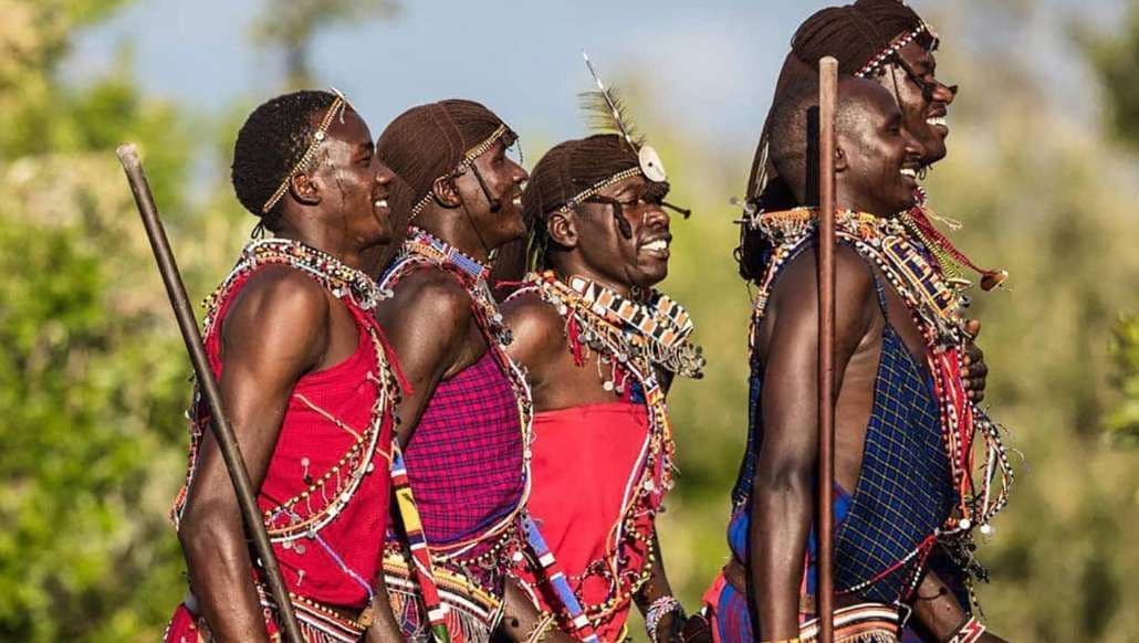 Un grupo de hombres con ropa tradicional colorida y collares de cuentas posan en fila, sonriendo al aire libre.