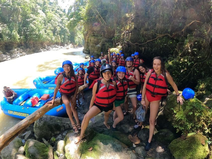 Un grupo WeRoad, con chalecos salvavidas rojos y cascos azules, posa para una foto en la orilla rocosa de un río junto a sus balsas.