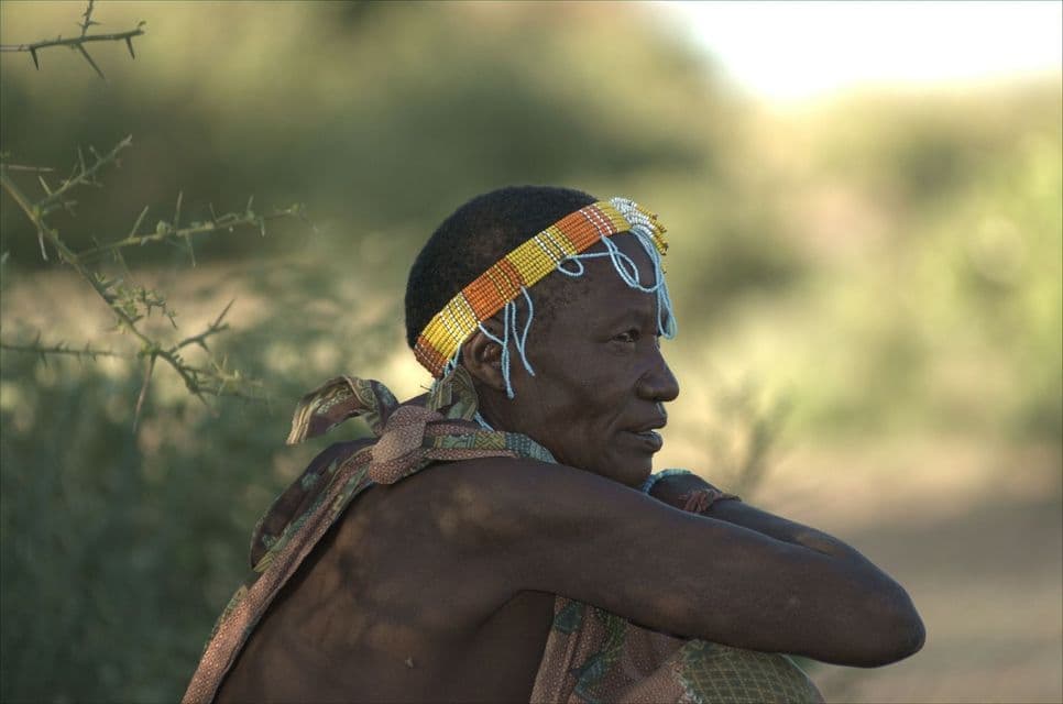 A person in profile wearing a colorful beaded headband and a patterned cloth, resting outdoors with a blurred background.