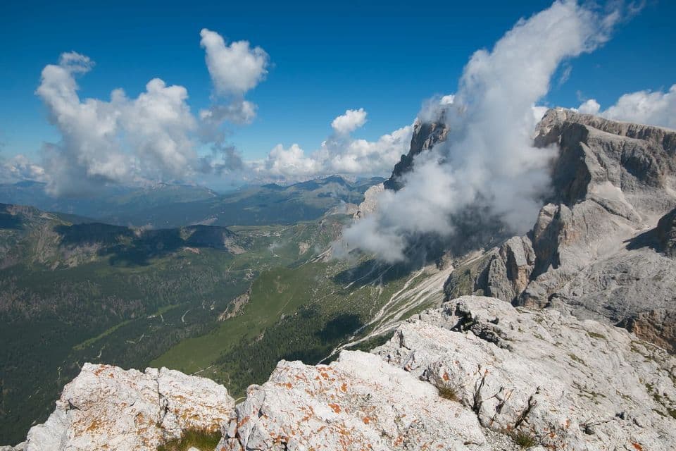 A view from a rocky mountain summit overlooking a vast green valley, with clouds swirling around a neighboring jagged peak.