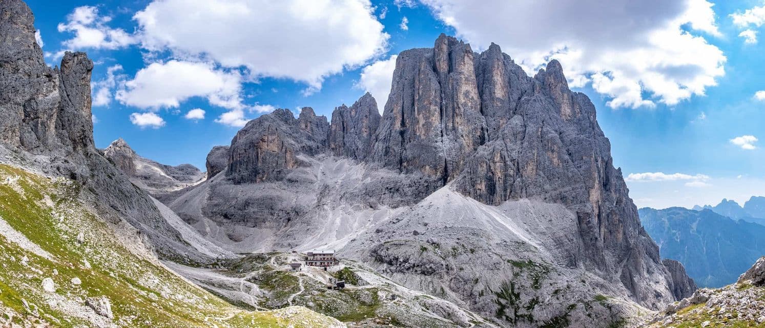 A mountain refuge is nestled in a valley below jagged rocky peaks under a partly cloudy blue sky.