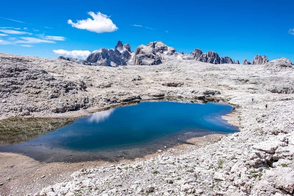 A small alpine lake reflects a blue sky in a vast, rocky landscape, with jagged mountain peaks rising in the distance.