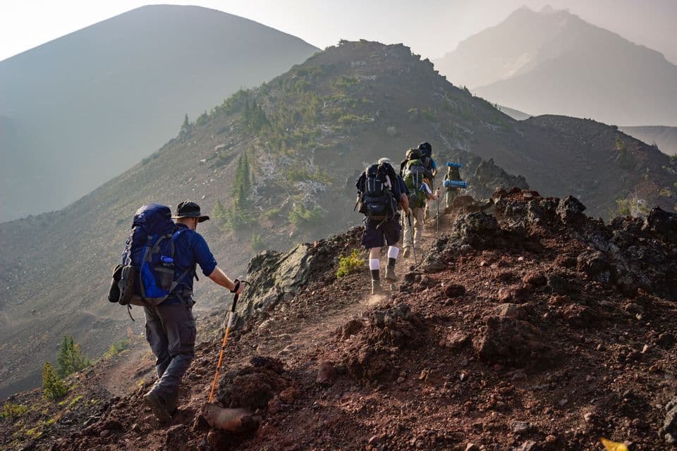 A WeRoad group trip hiking in single file up a narrow, rocky path on a mountainside with hazy peaks in the distance.