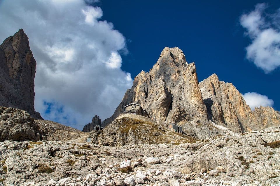 A stone refuge sits on a rocky outcrop at the base of jagged mountain peaks under a blue sky with white clouds.