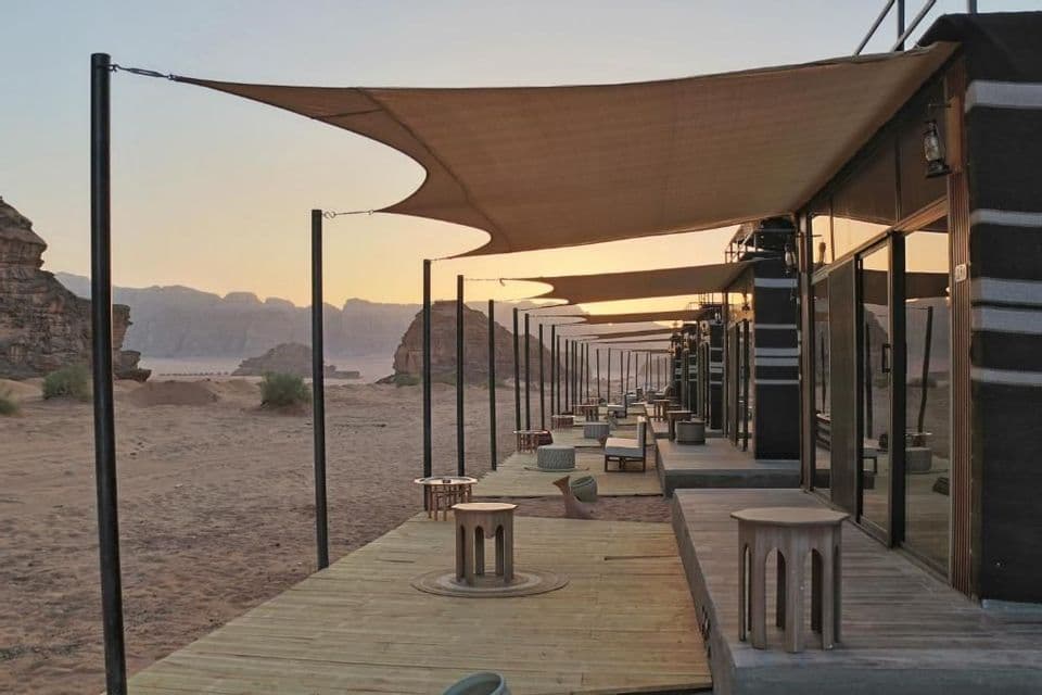 Cabine moderne nel deserto disposte lungo una piattaforma in legno con sedute, sullo sfondo di montagne rocciose al tramonto.