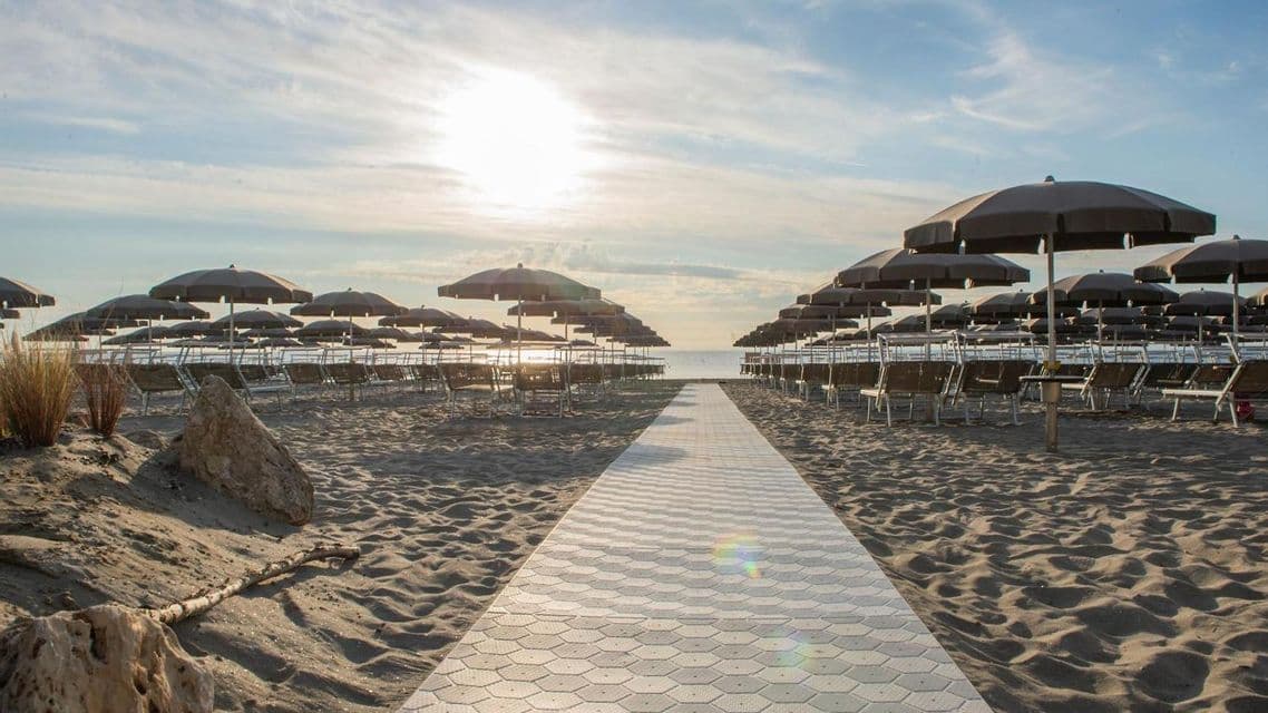 Una passerella piastrellata su una spiaggia sabbiosa conduce tra file di sedie e ombrelloni vuoti verso il mare al tramonto.