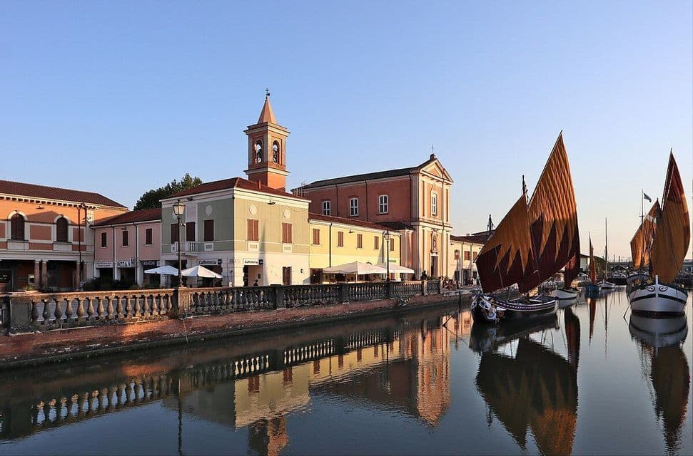 Barche a vela tradizionali con vele arancioni e marroni ormeggiate in un canale, con i loro riflessi e gli edifici colorati adiacenti visibili nell'acqua.