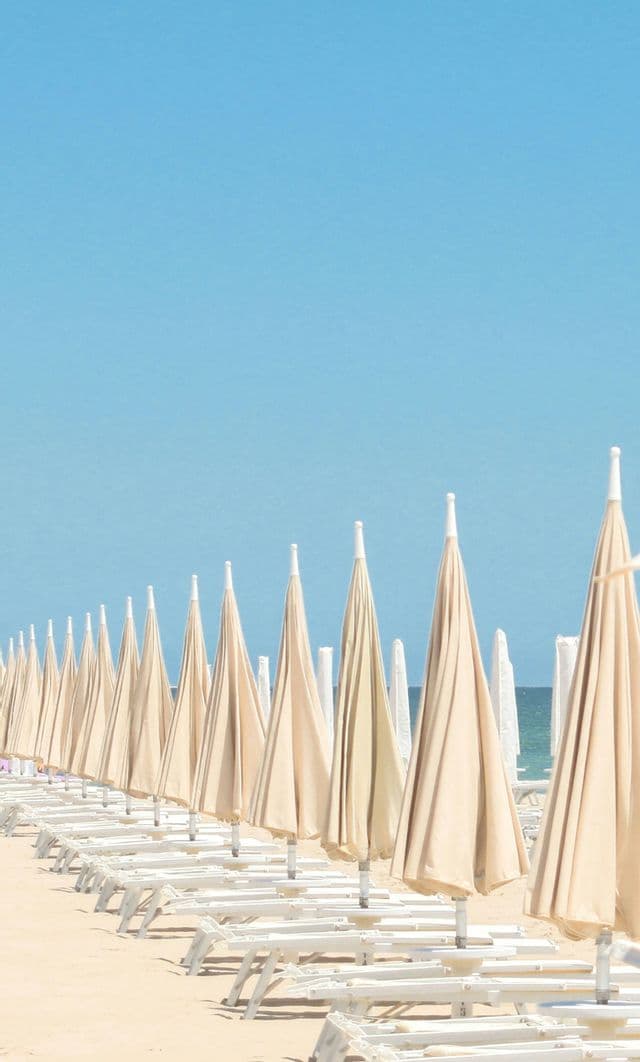 Fila di ombrelloni beige chiusi e lettini bianchi vuoti lungo una spiaggia sabbiosa sotto un cielo blu chiaro.