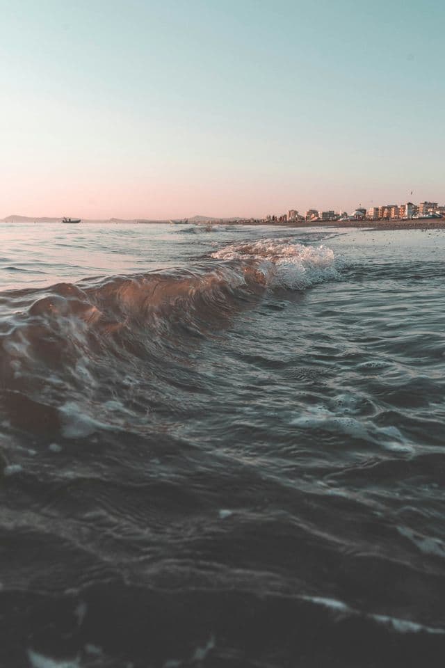 Primo piano di una piccola onda del mare che si infrange su una spiaggia, con lo skyline di una città visibile al tramonto.
