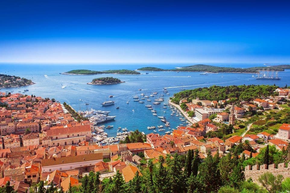 Blick aus der Vogelperspektive auf eine Küstenstadt mit Terrakottadächern, die einen Hafen voller Yachten und Boote überblickt, mit Inseln in der Ferne.