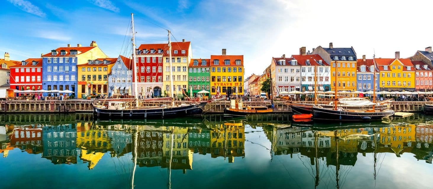 Une rangée de bâtiments colorés et de bateaux amarrés borde un canal, leurs reflets se mirant dans l'eau calme sous un ciel bleu.