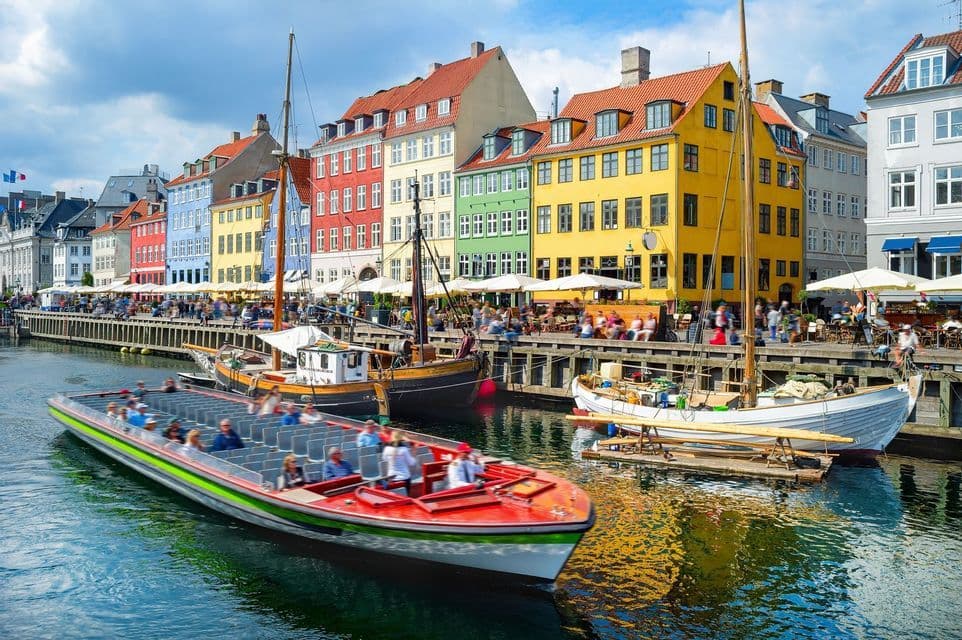 Un barco turístico lleno de gente navega por un canal bordeado de edificios coloridos y cafés frente al agua bajo un cielo azul.