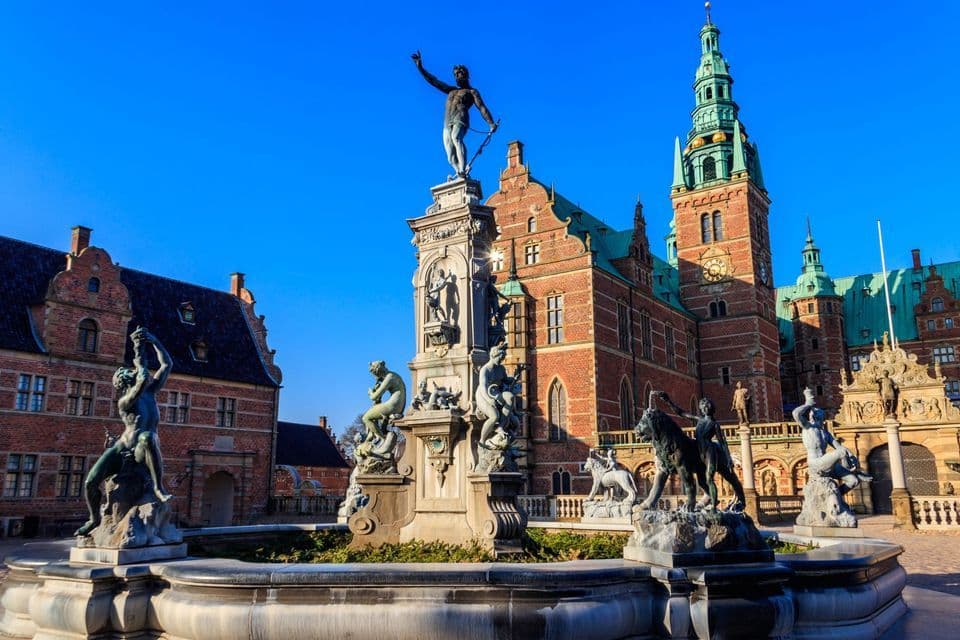 Ein kunstvoller Brunnen mit Bronzestatuen befindet sich auf einem Platz vor einem historischen Backsteingebäude mit Uhrenturm.