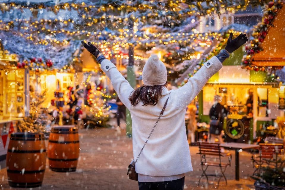 Una donna vista di spalle alza le braccia in un mercatino di Natale illuminato mentre la neve cade intorno a lei.