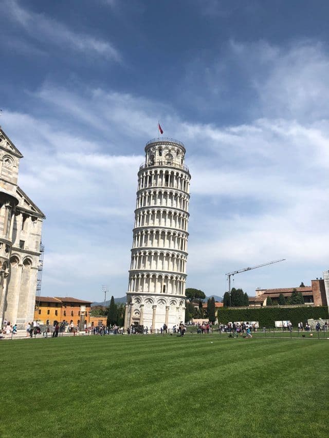 Der Schiefe Turm von Pisa steht auf einer großen grünen Wiese, mit Touristen an seinem Sockel unter blauem Himmel mit weißen Wolken.