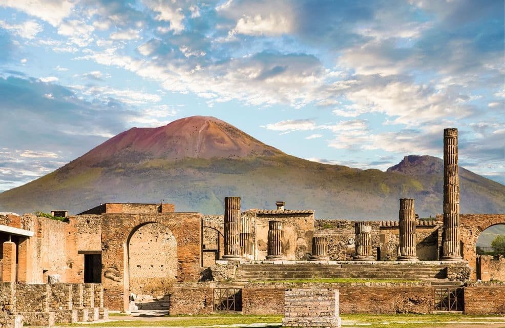 D'anciennes ruines avec des colonnes et des arches en pierre se dressent devant un grand volcan sous un ciel partiellement nuageux.