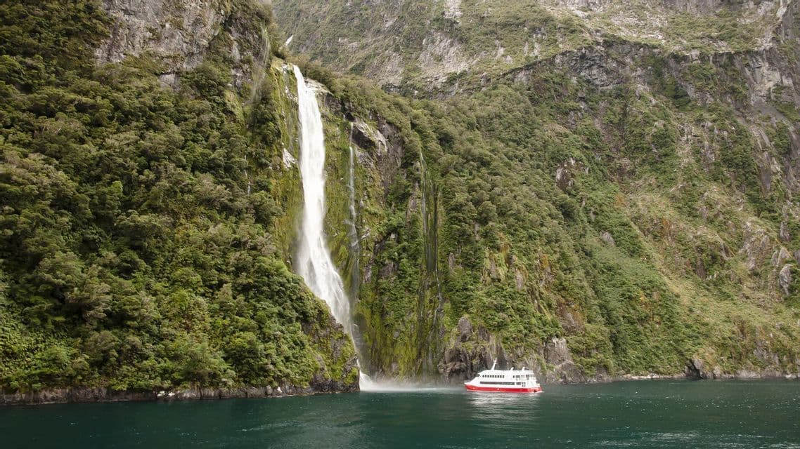 Una barca turistica rossa e bianca galleggia sull'acqua vicino alla base di un'enorme cascata che si riversa da una scogliera verde e rocciosa.