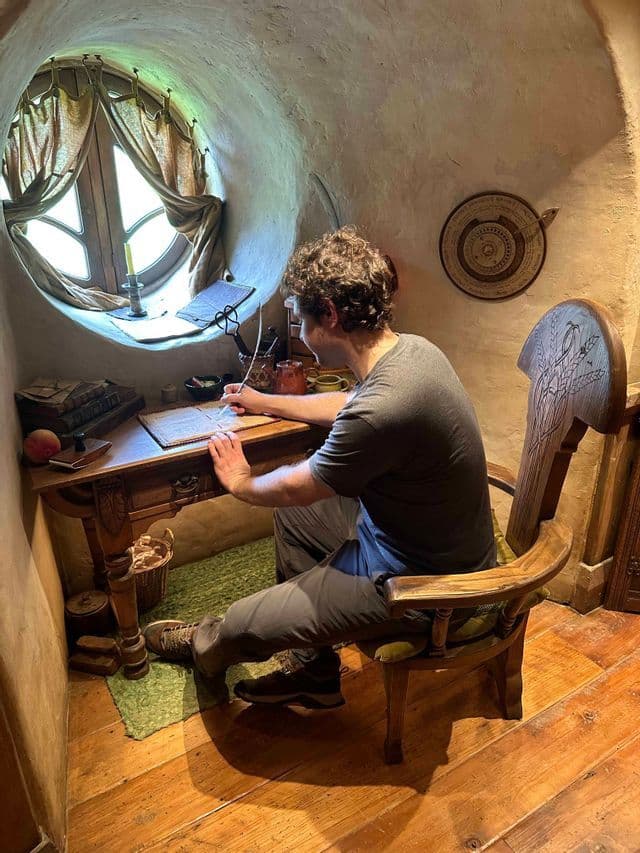 Un uomo è seduto a una scrivania di legno scrivendo con una penna d'oca accanto a una finestra rotonda in una stanza con pareti rustiche e curve.