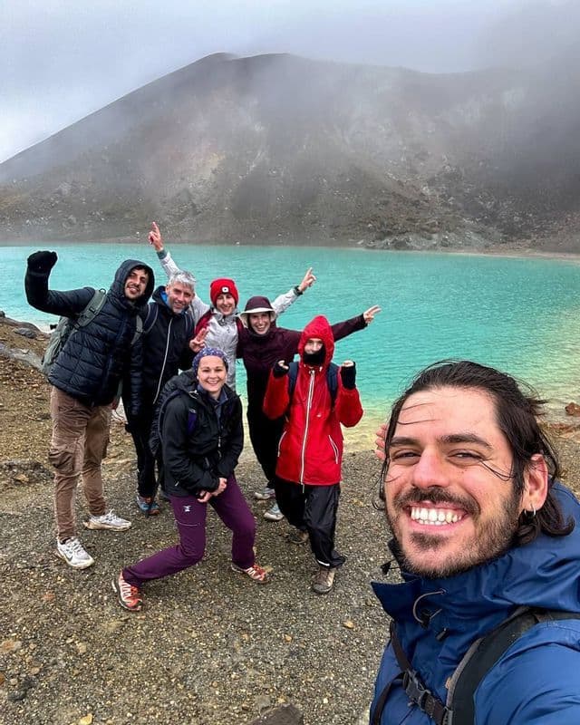 Un gruppo WeRoad si fa un selfie sulla riva di un lago turchese, con una montagna nebbiosa sullo sfondo.