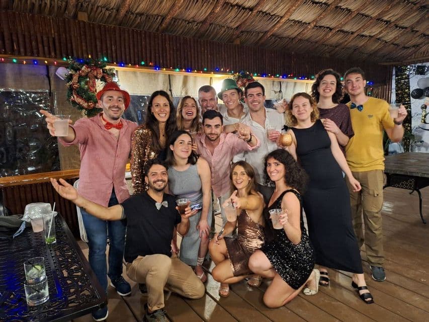 Un folto gruppo WeRoad in viaggio posa per una foto con drink e cappelli festivi durante una festa sotto un tetto di paglia.