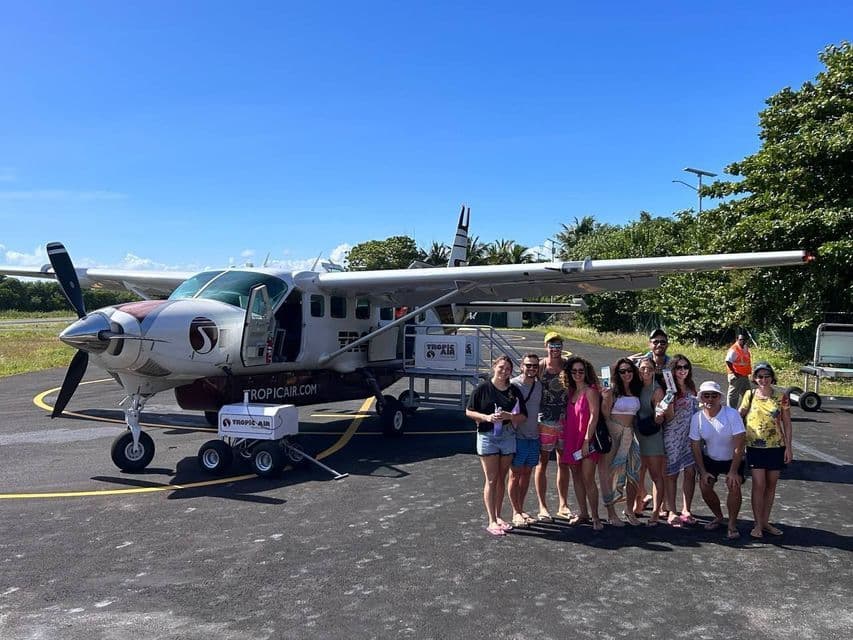 Un gruppo WeRoad posa per una foto sul piazzale di un aeroporto davanti a un piccolo aereo a elica di Tropic Air.